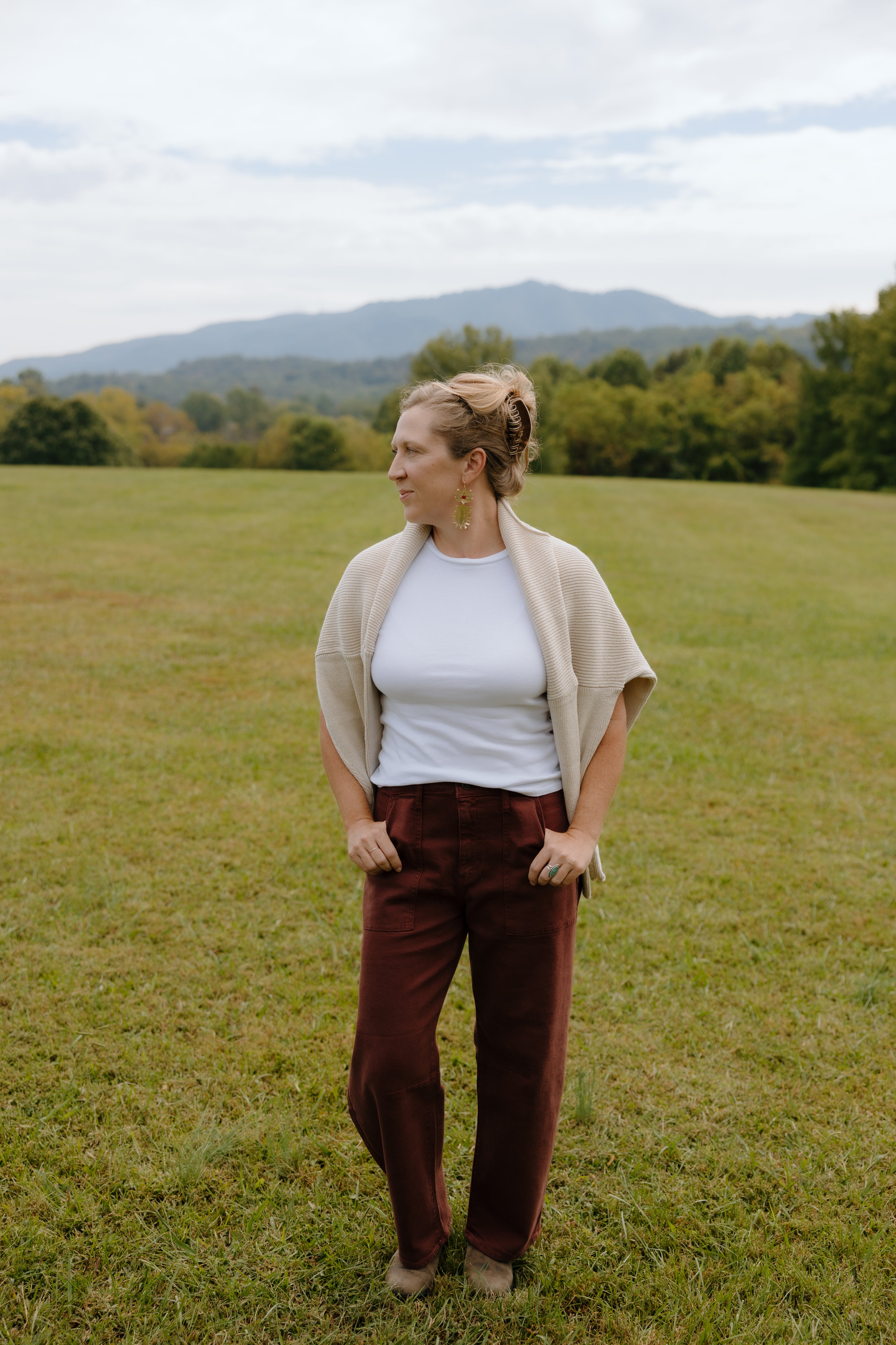 Woman standing in a grassy field wearing a white shirt, brown pants, and a beige cardigan.