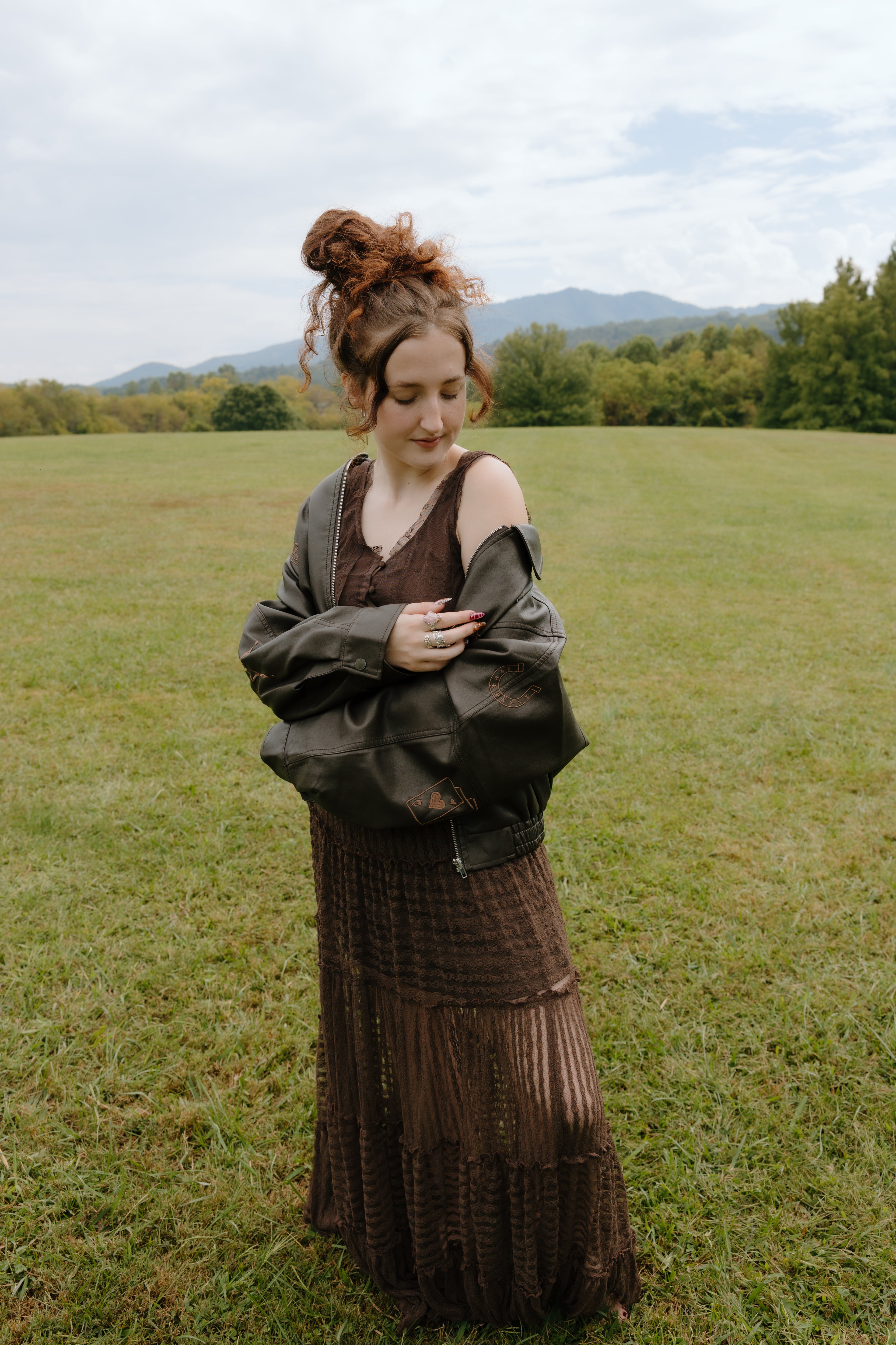 Woman in a brown dress standing in a grassy field with mountains in the background